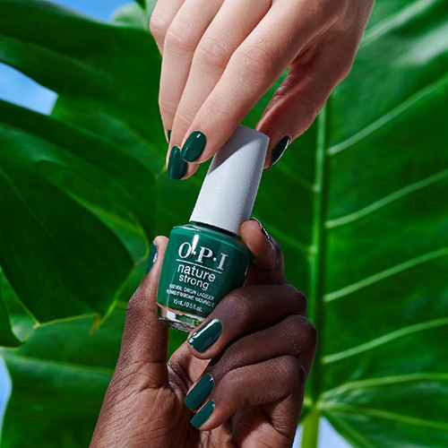 Two hands holding a green OPI Leaf by Example Color nail polish bottle in front of a big green leaf