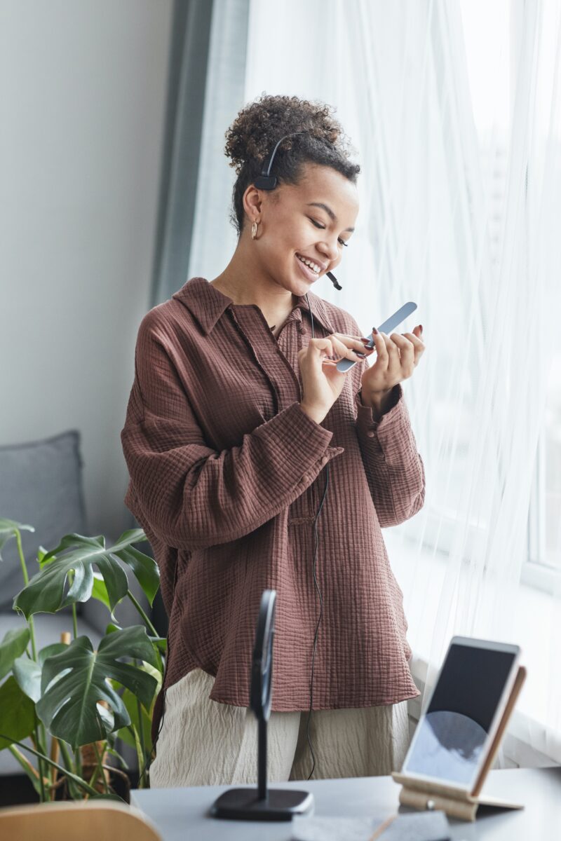 A beautiful, tall woman wearing a brown shirt and beige pants with painted red nails smiles while using a nail file to shape her nails.