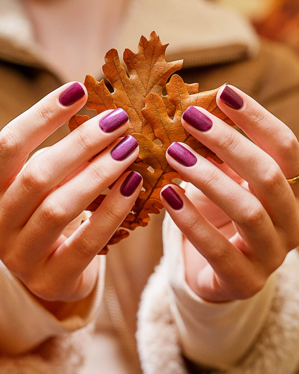 A faceless woman wearing a white shirt is holding an autumn leaf with fuchsia berry painted nails using one of the Change Of Pace collection shades from Gelish for Fall 2023