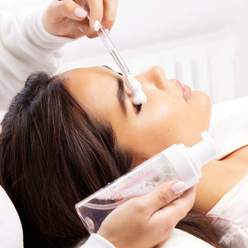A beautiful woman is lying on a beauty salon bed while a lash technician applies cleanser from the JB Lashes brand to her natural lashes.