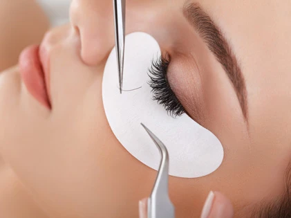 A faceless lash technician wields a silver pair of specialized eyelash tongs, skillfully applying extension lashes to the client's natural eyes.