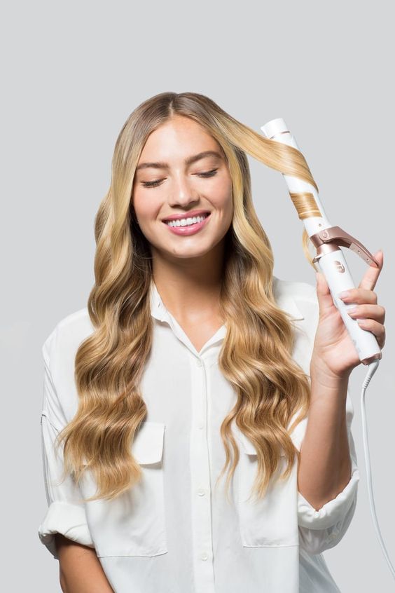 A beautiful smiling woman with long blond wavy hair is holding a hair curling iron and wearing a white shirt.