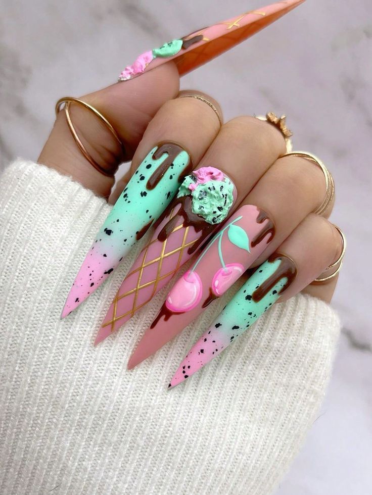 A woman's hand is adorned with pink and green painted nails, each featuring intricate ice cream, chocolate, and cherry designs. Nail art trends