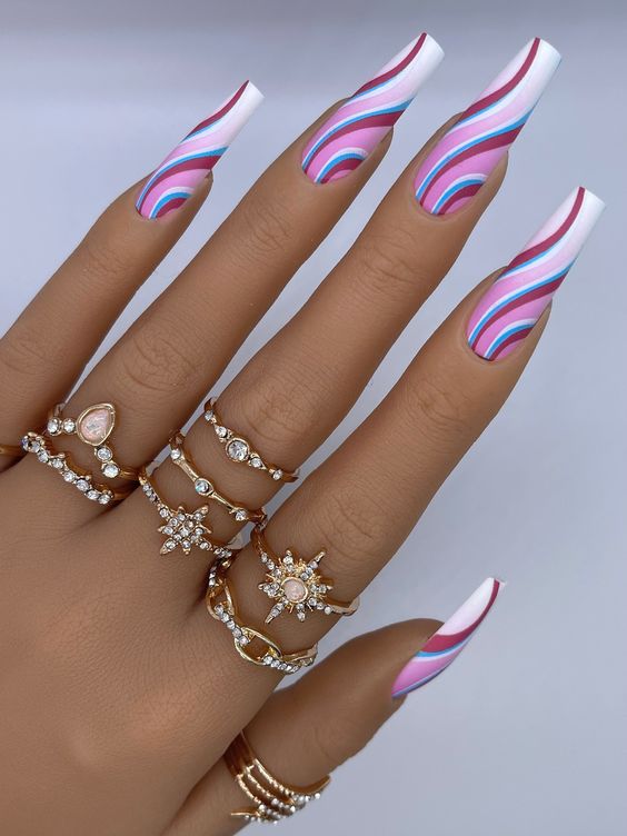 A woman's hand, adorned with gold rings, showcases painted nails in shades of blue, purple, white, and light pink, featuring a Barbie wavy nail design. Nail art trends