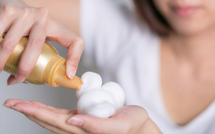 A woman in a crisp white t-shirt holds a bottle of hair mousse, applying the product onto her hand before distributing it through her hair.