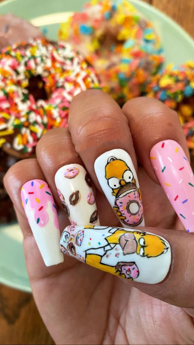 A woman's hand is adorned with pink and white painted nails, each featuring intricate designs. Among them are donut designs and a yellow Simpson character wearing a white shirt and holding a donut. Spring aail art trends