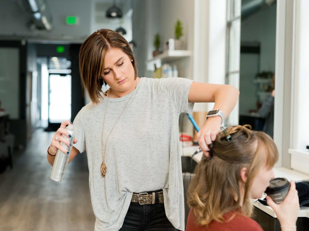 A hair professional wearing a gray shirt and an Apple Watch is holding a spry strand and applying it to the client's hair.