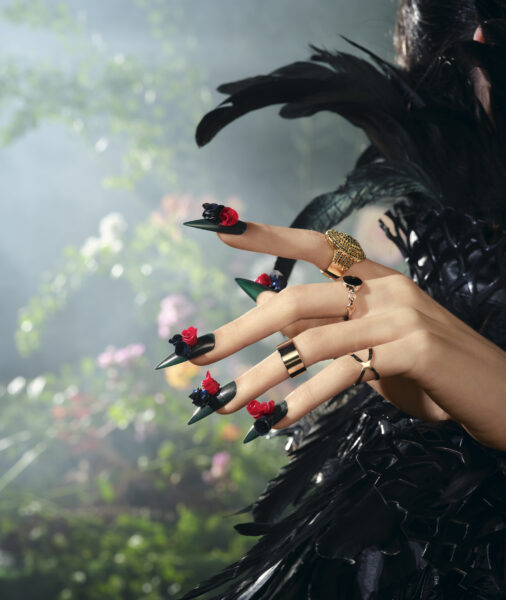 A faceless woman wearing a black dress adorned with feathers, gold rings on her fingers, and bold green nail polish from the new CND Winter 2023 collection, the Magical Botany. Her nails were further embellished with intricate black and red floral nail designs.