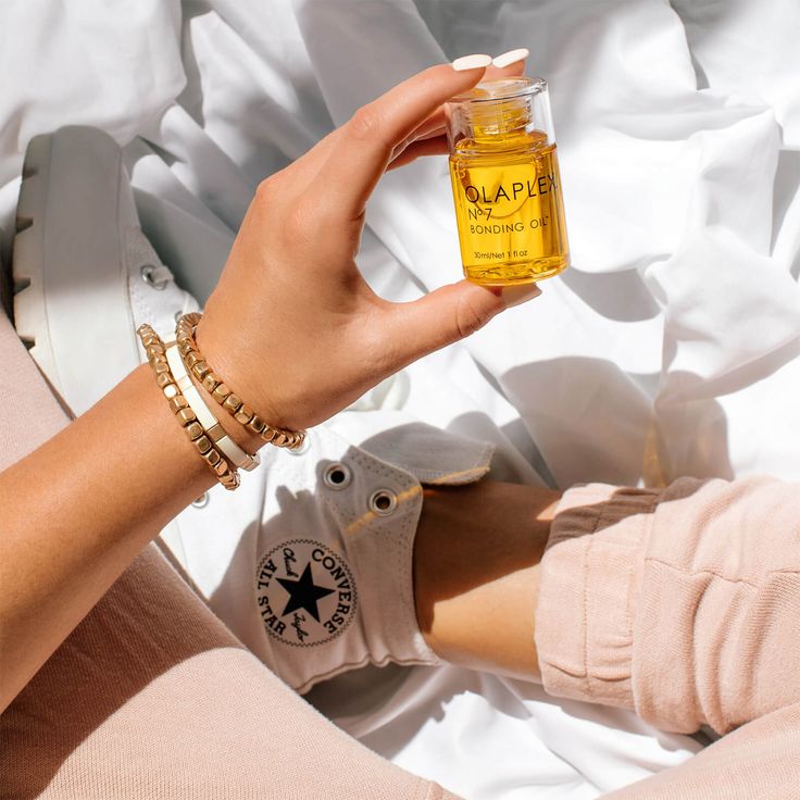 A faceless woman wearing boots holding a bottle of Olaplex oil on a bed.