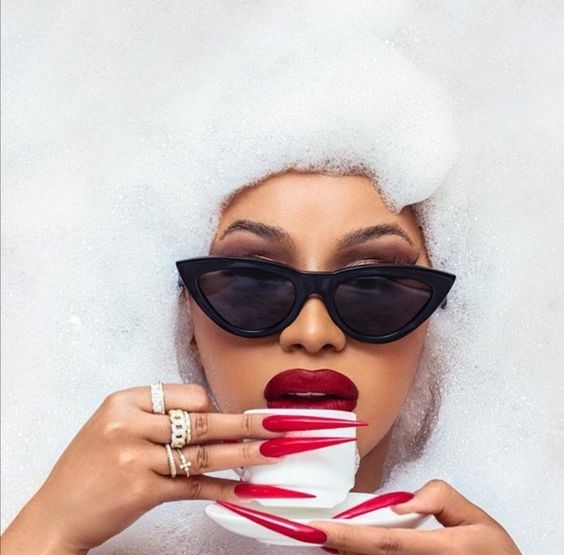 Cardi B wearing sunglasses and red nail polish is holding a cup of coffee.
