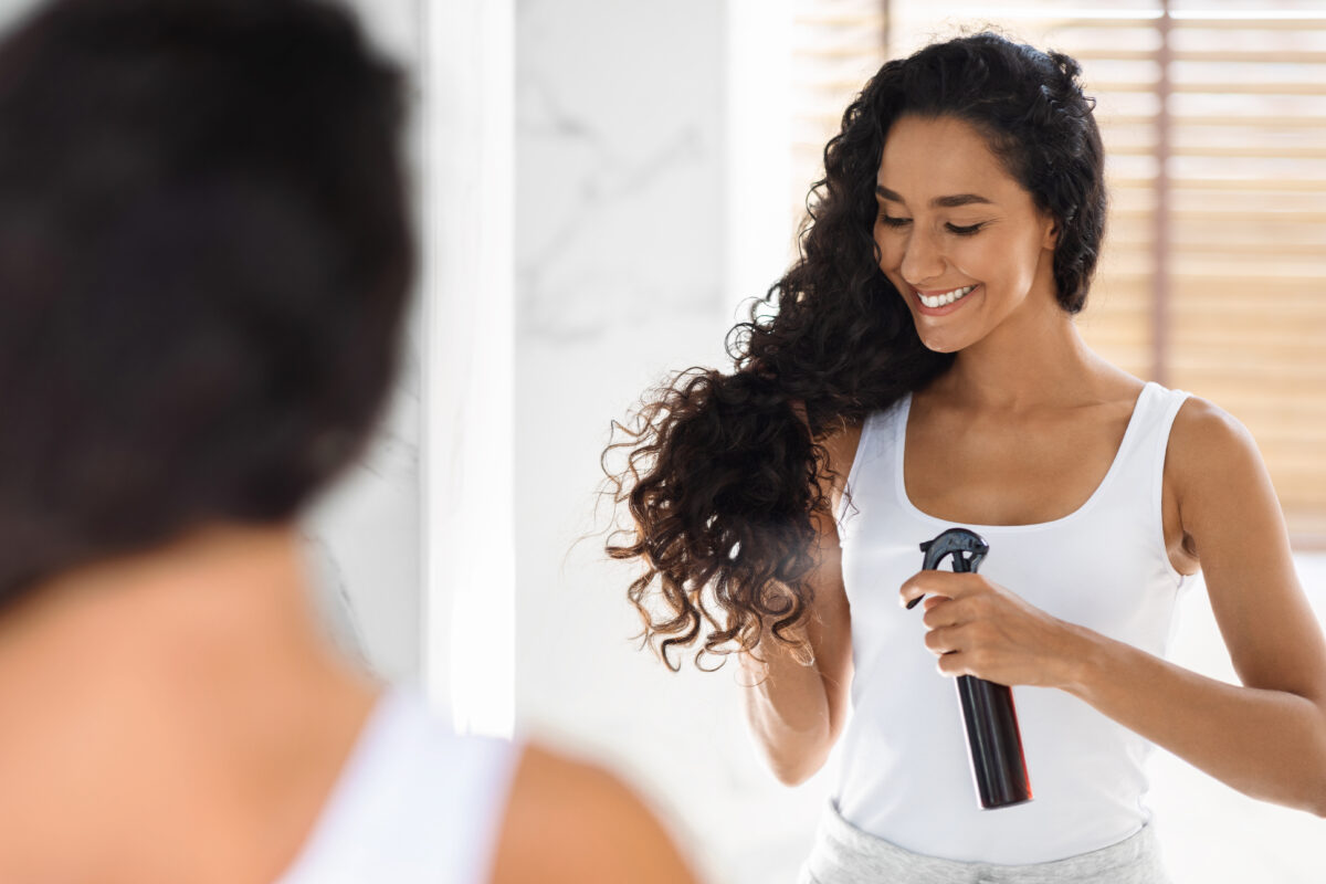 Beautiful Young Lady Applying Hair Spray For Curls While Standing Near Mirror