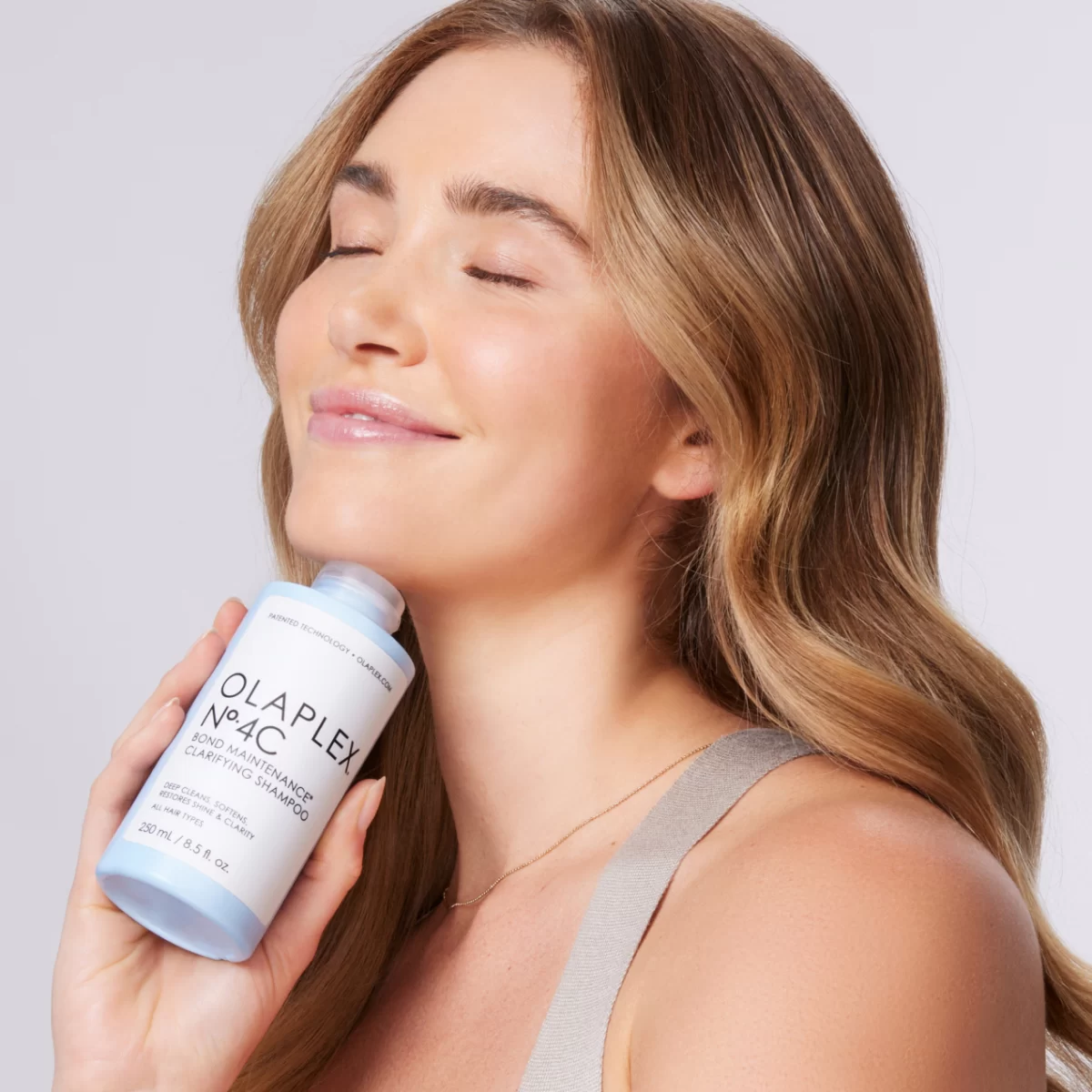 A beautiful blonde woman smiles while holding the Olaplex shampoo.