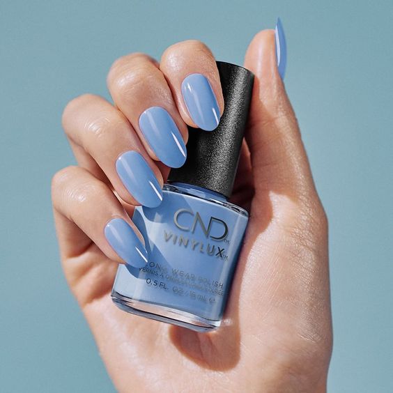 A faceless woman holding a CND Vinylux nail bottle in light blue nail shade, with her nails painted in the same color, against a blue background