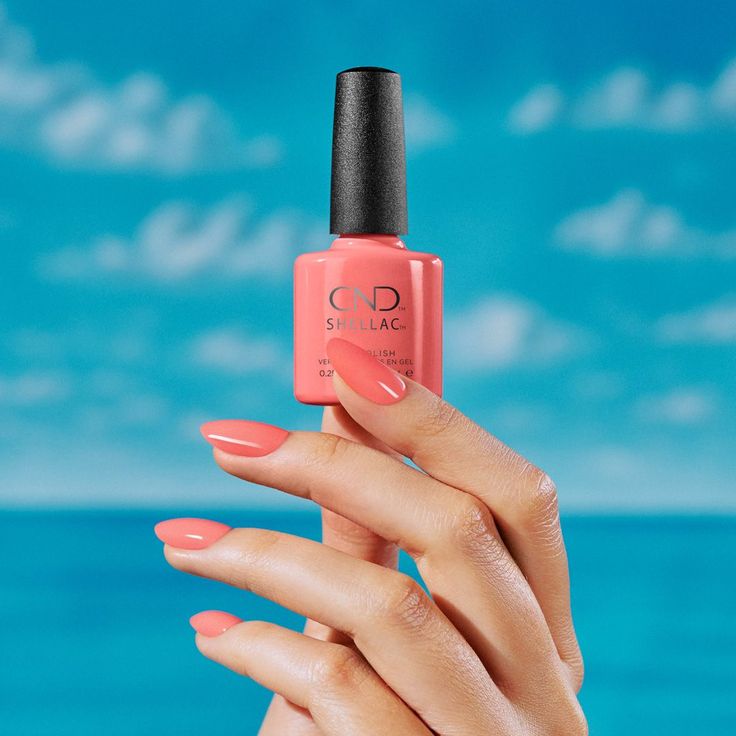 A faceless woman holding a CND Shellac nail bottle in a pink shade, with her nails painted in the same color, against a backdrop of a blue sea and sky.