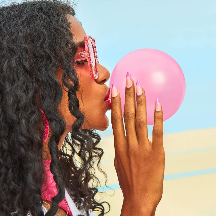 A beautiful woman is at the beach wearing pink glasses and earrings with long painted nails in the pink shade from the Summer Make The Rule collection from OPI's summer 2023 collection. Summer Make the Rules - OPI Summer 2023 Collection