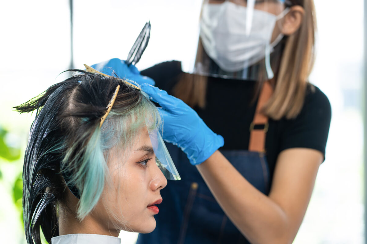 Professional hairstylist dyeing young woman customer's hair in salon. Hairdresser wear face shield and mask to prevent from coronavirus infection, give service treatment to woman in beauty barber shop