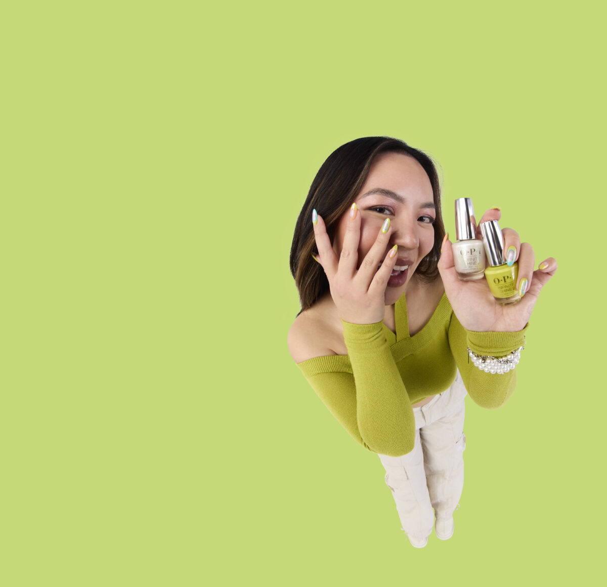 A girl wearing a green top and white pants holding up two OPI Spring 2024 nail polish bottles in green and yellow shades against a green background.