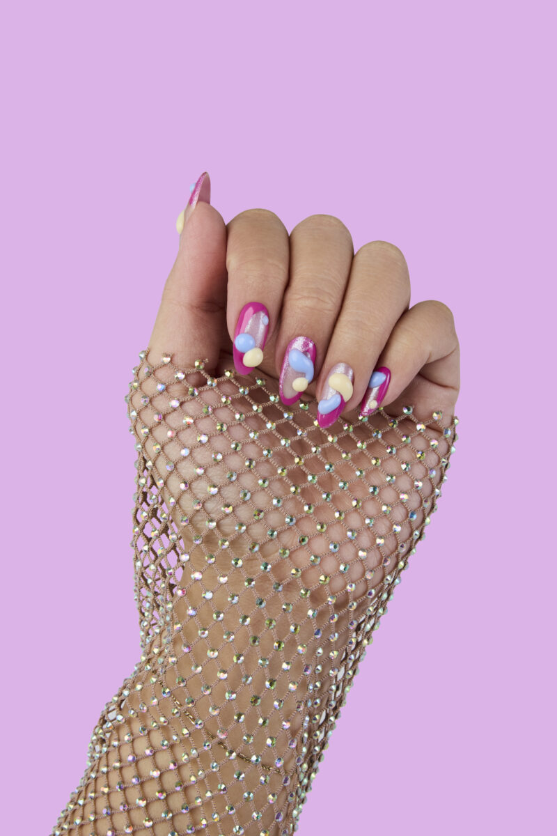 A hand with pink nails and a mesh glittery glove against a pink background. The nails have artistic designs using the OPI Spring 2024 collection.