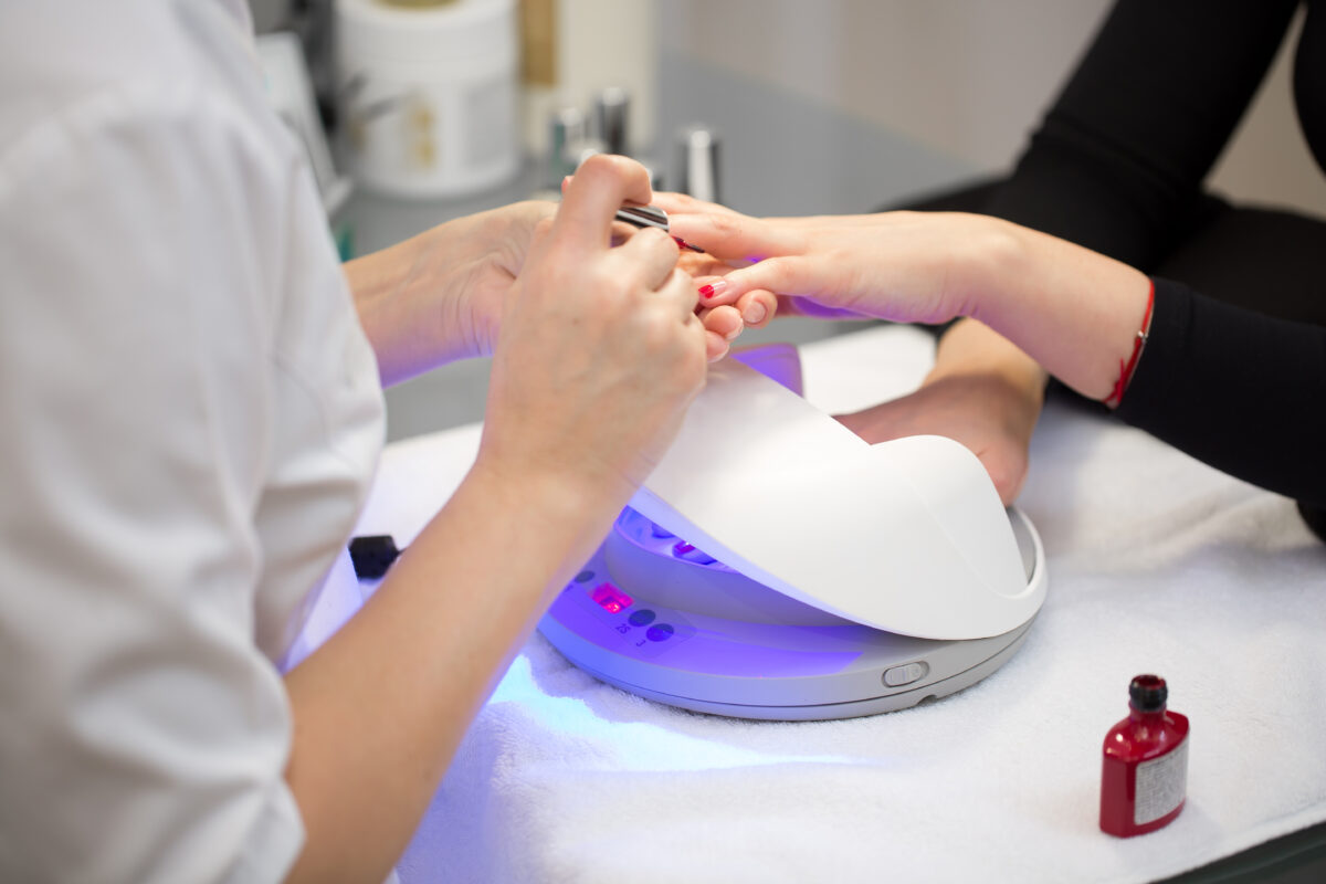 Close-up of the hand of a manicurist who applies red gel Polish on the nails of a young girl. Girl dries gel nail Polish in UV lamp.