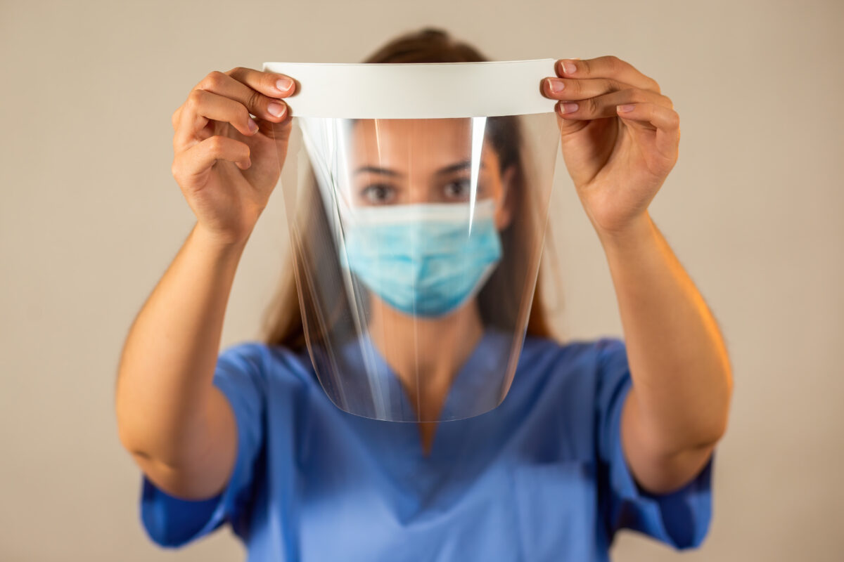 Female professional in blue shirt holding protective face shield in front of her. Concept of safety during working. Young woman with brown hair using protection against viruses.