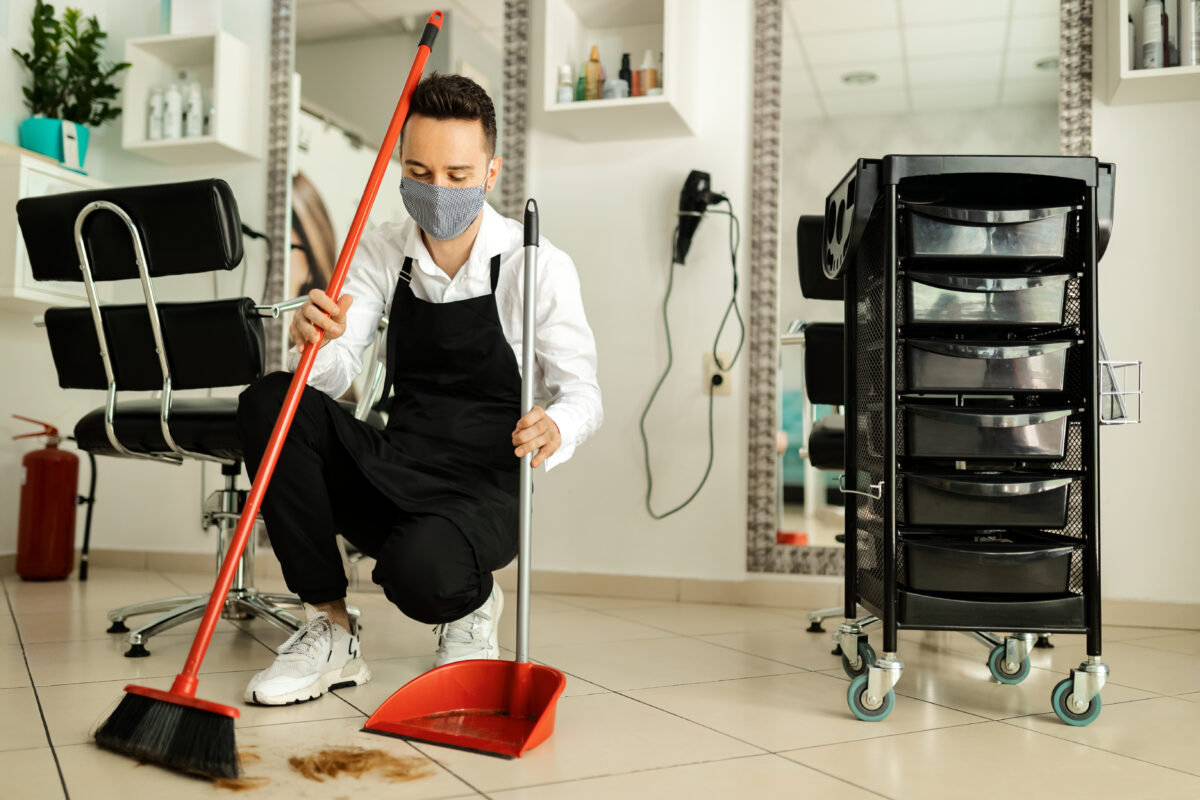Hairstylist with protective face mask cleaning salon's floor with a broom after a haircut.
