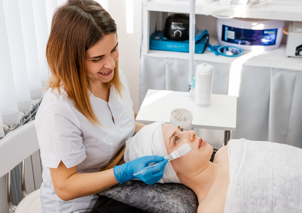 Hands of cosmetology specialist applying gold facial mask using brush, making skin hydrated and face glowing and skin. Attractive brunette relaxing with closed eyes and enjoying spa procedures.