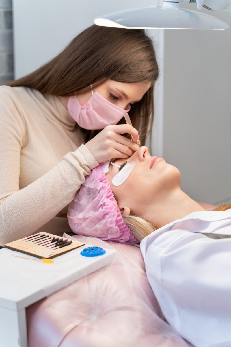 Eyelash extension master with tweezers in his hands performs eyelash correction close-up. Girl's eye with black artificial eyelashes closeup. Eyelash extension procedure in beauty salon. Lashmaker at work. Concept spa lash.