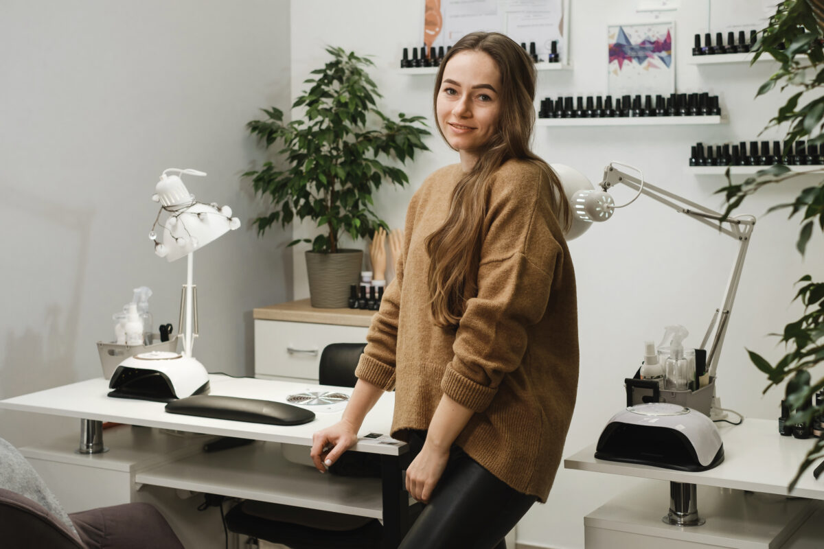 Nail service master. Portrait of professional young brunette sitting at modern nail salon workplace and smiling while waiting client. Beauty service Occupation concept