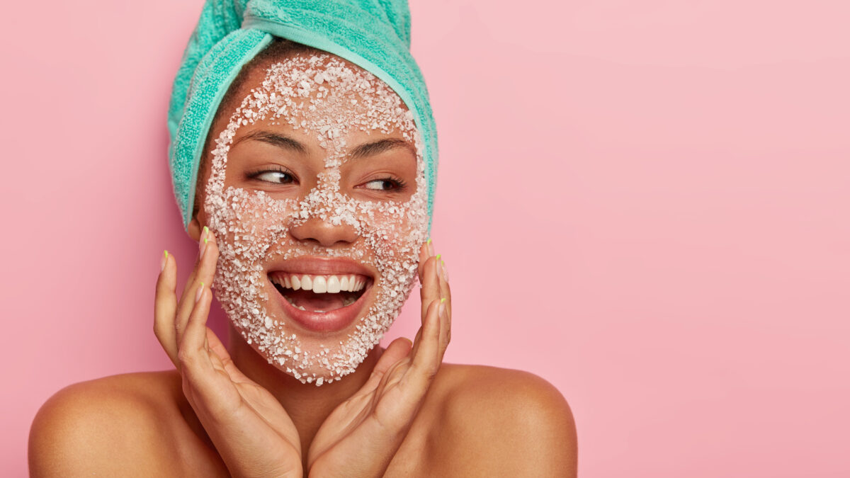 Portrait of happy cheerful woman improves condition of face skin, applies peeling mask, wants to have pleasant appearance, shows bare shoulders, being in high spirit, models indoor. Spa treatment