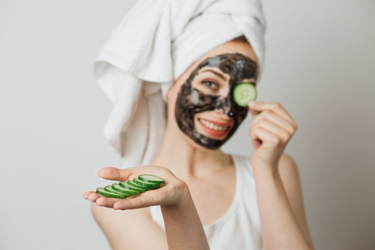 Portrait of smiling young woman with cosmetic mask on face holding slice of cucumber near eye. Isolated over grey studio background.