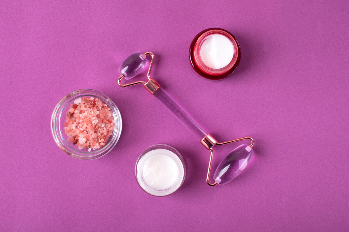 Skincare set with jade roller for facial massage, pink salt scrub and moisturizer cream on purple background. Beauty treatment flat lay
