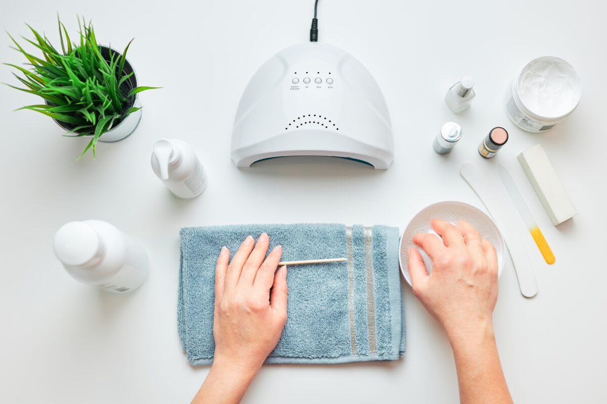 Woman preparing nails to apply gel hybrid polish using UV lamp. Beauty wellness spa treatment concept. Cosmetic products, UV lamp, green leaves on white table. Spa, manicure, skin care concept. Flat lay, overhead view