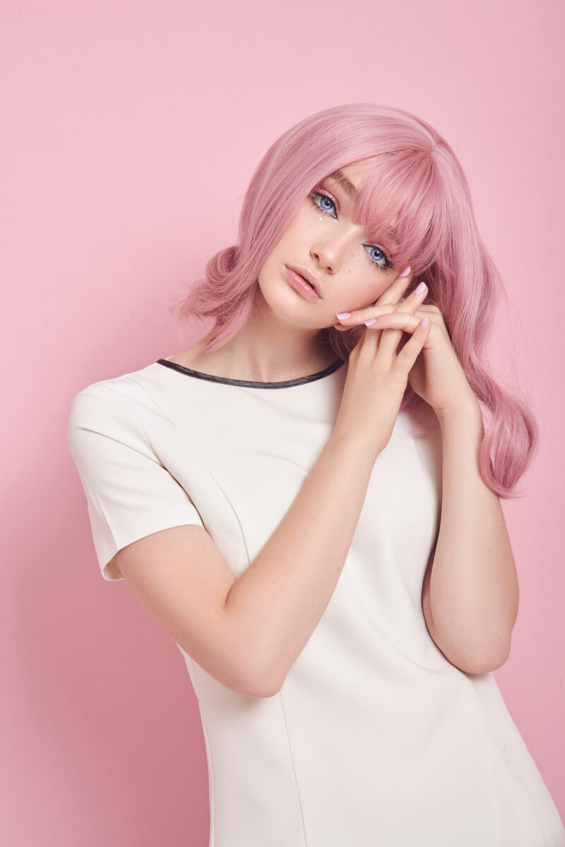 A model with pink hair, wearing a white dress poses against a pink background, showcasing the vibrant colors of the Entity Spring 2024 Cutie Couture nail polish collection. The model’s nails are painted with shades of the collection. The image conveys a sense of sweetness and fun, reflecting the inspiration of the collection.