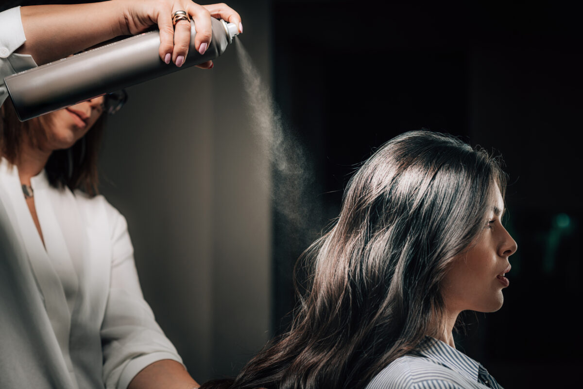 Hairdresser spraying woman’s long black hair with hairspray.