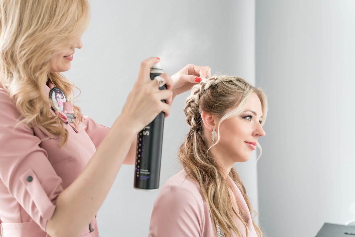 Hairdresser sprinkles hairspray on a client's hair. The girl sits in front of the mirror in the hairdresser's chair. Fixing the hairstyle with varnish. Professional hair styling in the salon. Two persons in the photo. A young woman of European appearance makes hair styling to a client. Beauty care. Soft focus.
