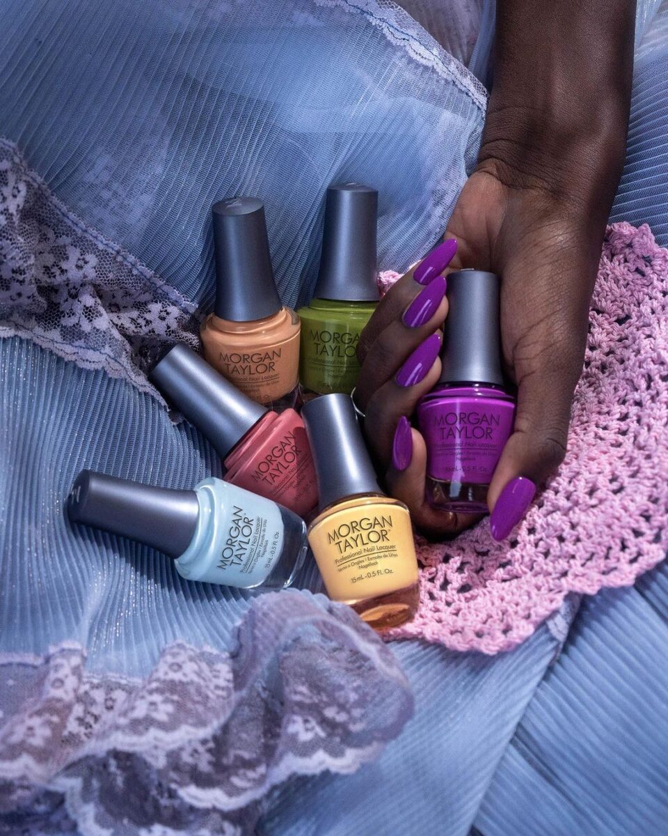 A faceless woman wearing purple nail polish on her nails holds a Morgan Taylor nail polish bottle, with other Morgan Taylor nail polish bottles beside her.