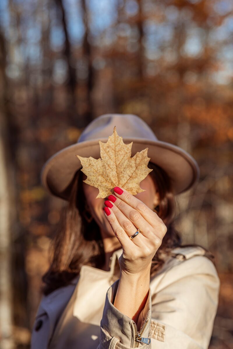 A woman hand elegantly displaying one of the Fall 2024 Nail Colors with a dried maple leaf, set against a soft-focus autumnal backdrop, highlighting the seasonal trend in nail fashion. Fall 2024 nail trends. Fall nails. 2024 nail trends
