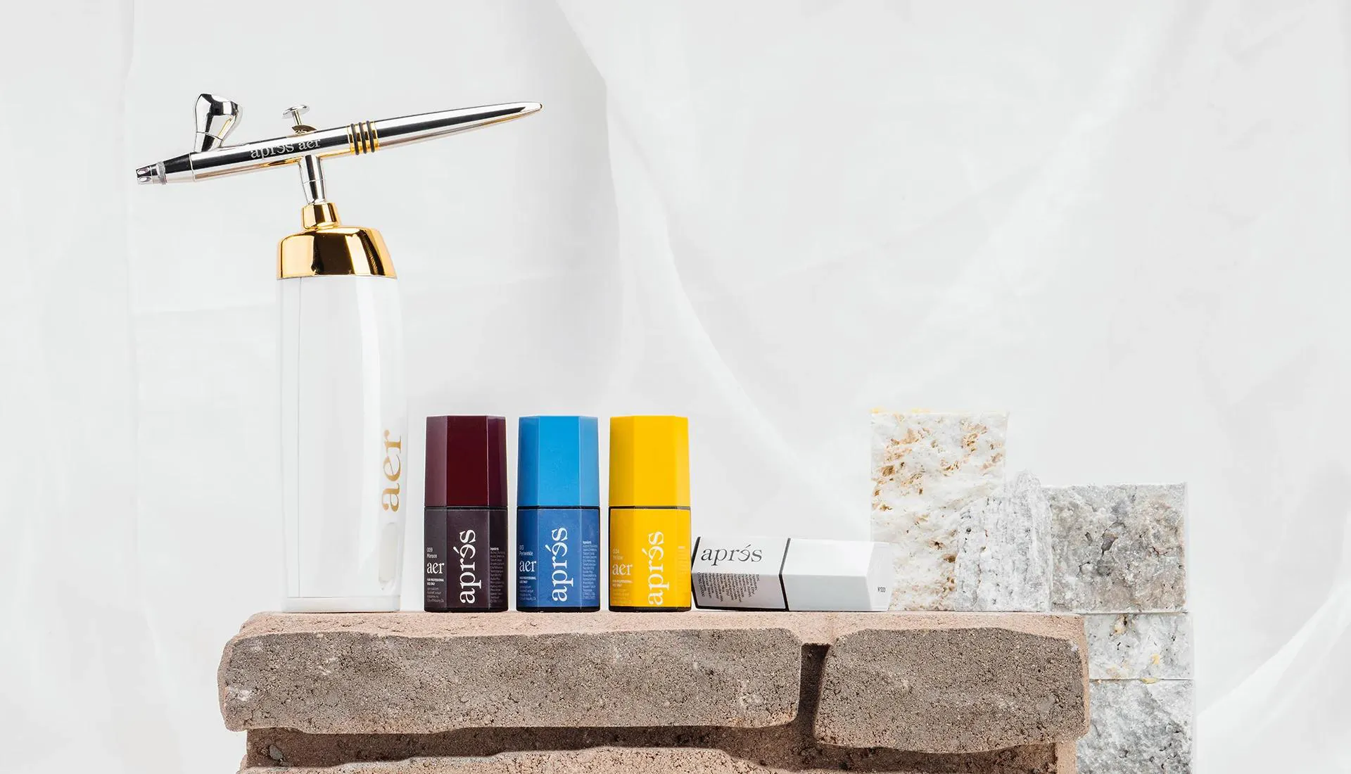 The newest Apres airbrush using new, cutting-edge gel technology. Nail Airbrush. gel polish. airbrush gel polish. A collection of nail care products displayed on multi-level marble and stone slabs. The arrangement includes a white nail airbrush with a gold and silver pump, labeled ‘Aprés. Next to it are three small, colorful bottles with black caps in blue, red and yellow, representing different colors of nail polish or gel. The products are showcased on two stone slabs, creating a layered effect. The elegant background features a light marble texture.