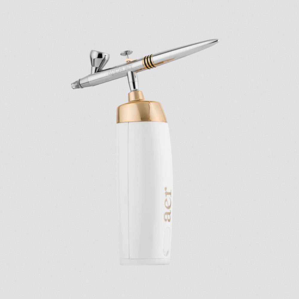 A sophisticated Apres Nail Brand airbrush tool, featuring a chic white body with elegant gold accents. The top part is metallic, likely chrome, with a slim nozzle and an attached transparent paint reservoir, highlighting its use for precise and artistic nail airbrush designs.