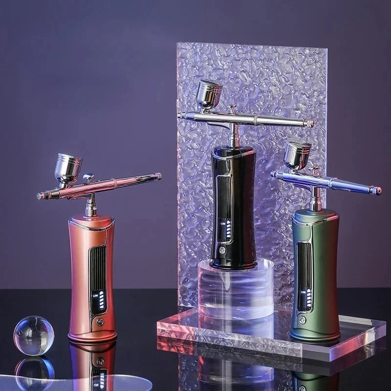 A trio of professional nail airbrush devices, each featuring a distinct color scheme: one in black with copper accents, another in metallic blue, and the third in green. They are elegantly displayed on transparent acrylic stands with a chrome finish support rod, against a backdrop of purple with a textured silver panel. The airbrushes boast a sleek and contemporary design, indicative of their precision and professional-grade capabilities in nail artistry. A decorative clear glass sphere is also present in the foreground, adding an artistic touch to the arrangement.
