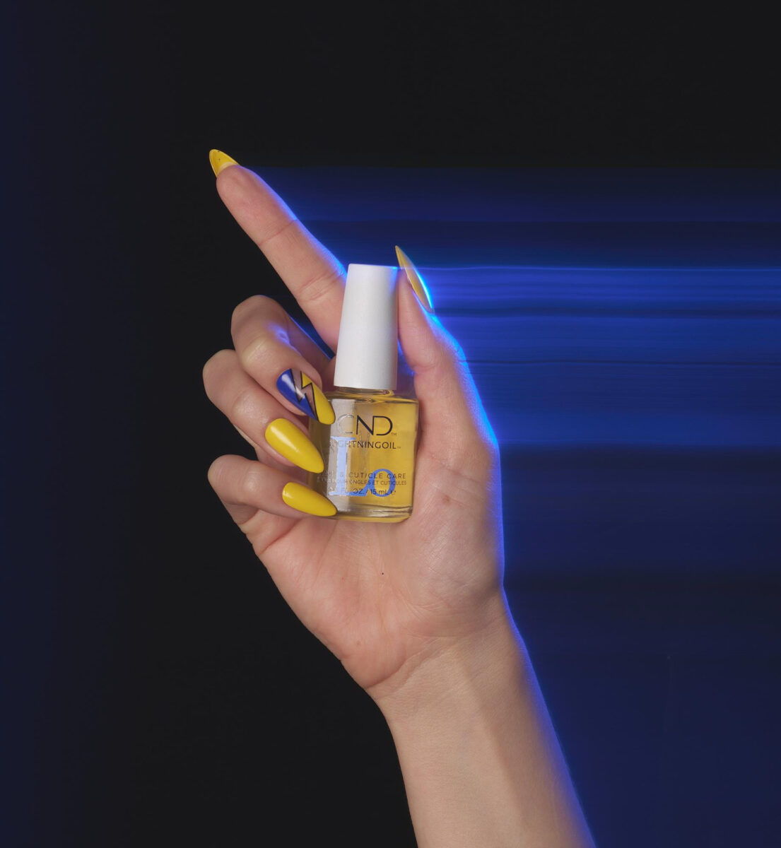 A faceless woman holding a bottle of the new CND LIGHTNINGOIL product, with her nails painted in bright yellow and blue nail polish featuring nail designs, against a blue background.