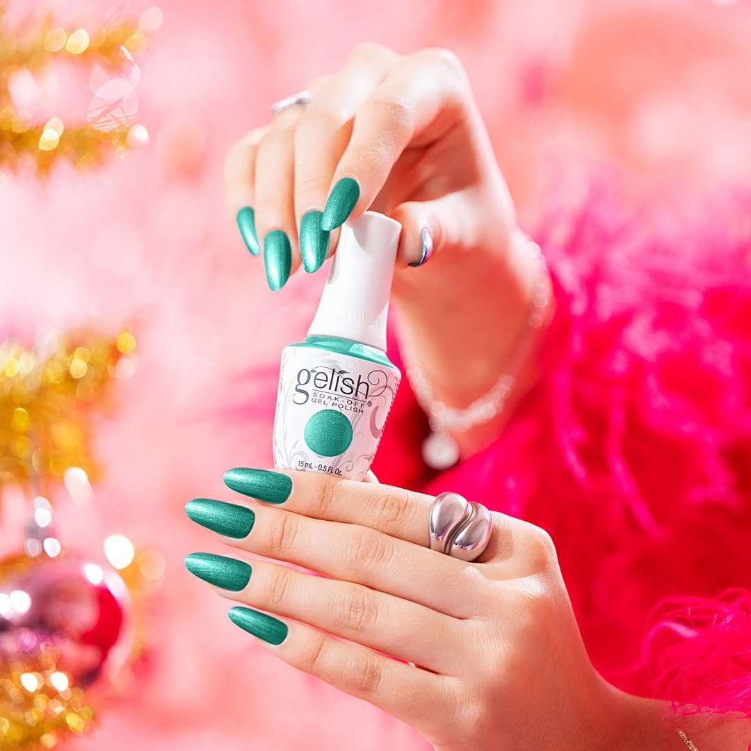 A close-up of hands holding a bottle of Gelish nail polish. The nails are painted in a shimmery green color that matches the nail polish bottle. The background is pink and blurred, adding a vibrant and colorful touch. The person is also wearing rings on their fingers, enhancing the aesthetic appeal of the image. This represents Gelish's Winter 2024 'A Fuzzy Feeling' collection.