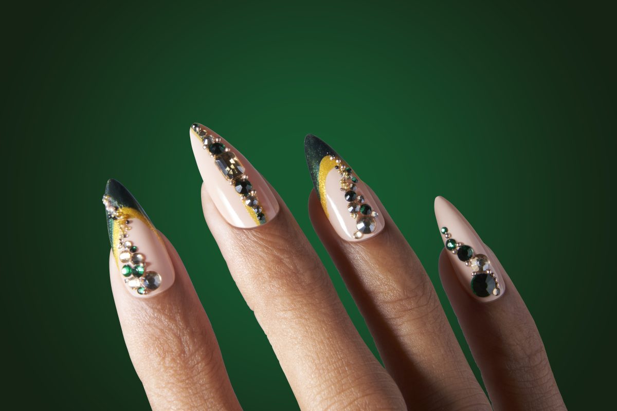 a close-up image showcasing a hand with nails painted in a variety of colors from the opi winter 2024 nail polish collection. the nail art features a holiday theme with intricate designs including dark green, gold glitter, and embellishments resembling tiny gemstones. the background is a solid green color, emphasizing the festive and luxurious feel of the nail designs.