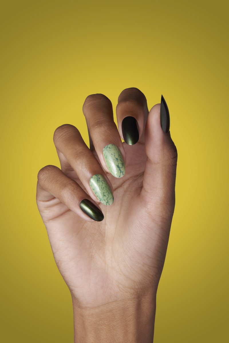 A close-up of a hand with a holiday-themed manicure from the OPI Winter 2024 Nail Polish Collection. The nails are long and feature a mix of shiny, metallic finishes and speckled marble patterns, set against a vibrant yellow background.