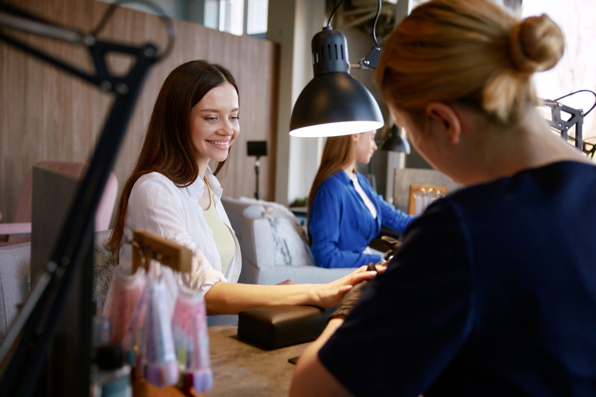 Happy laughing young woman friends getting manicure and gossiping together. Professional nail salon and satisfied clients. 