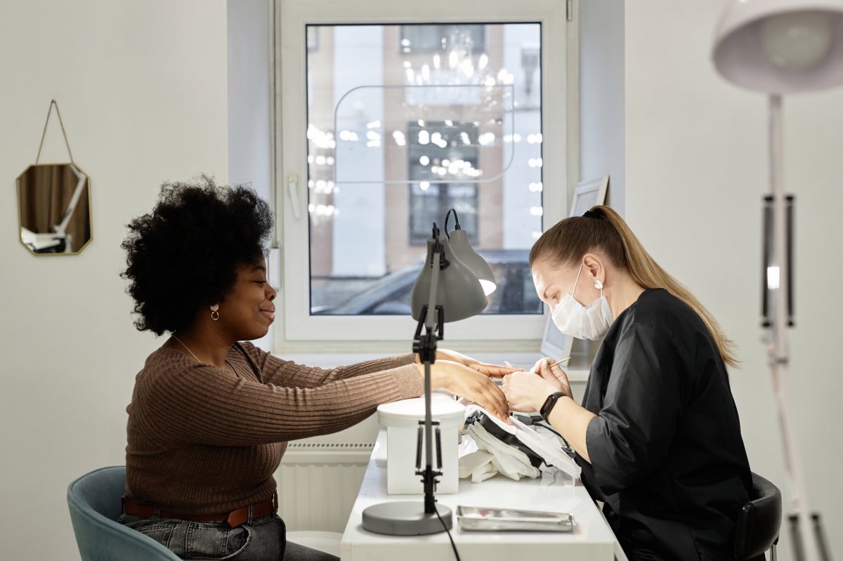Side view shot of female nail master working on manicure of happy African American client with curly hair. Black woman at nail tech desk getting nails cut, filed, and shaped, copy spaceز