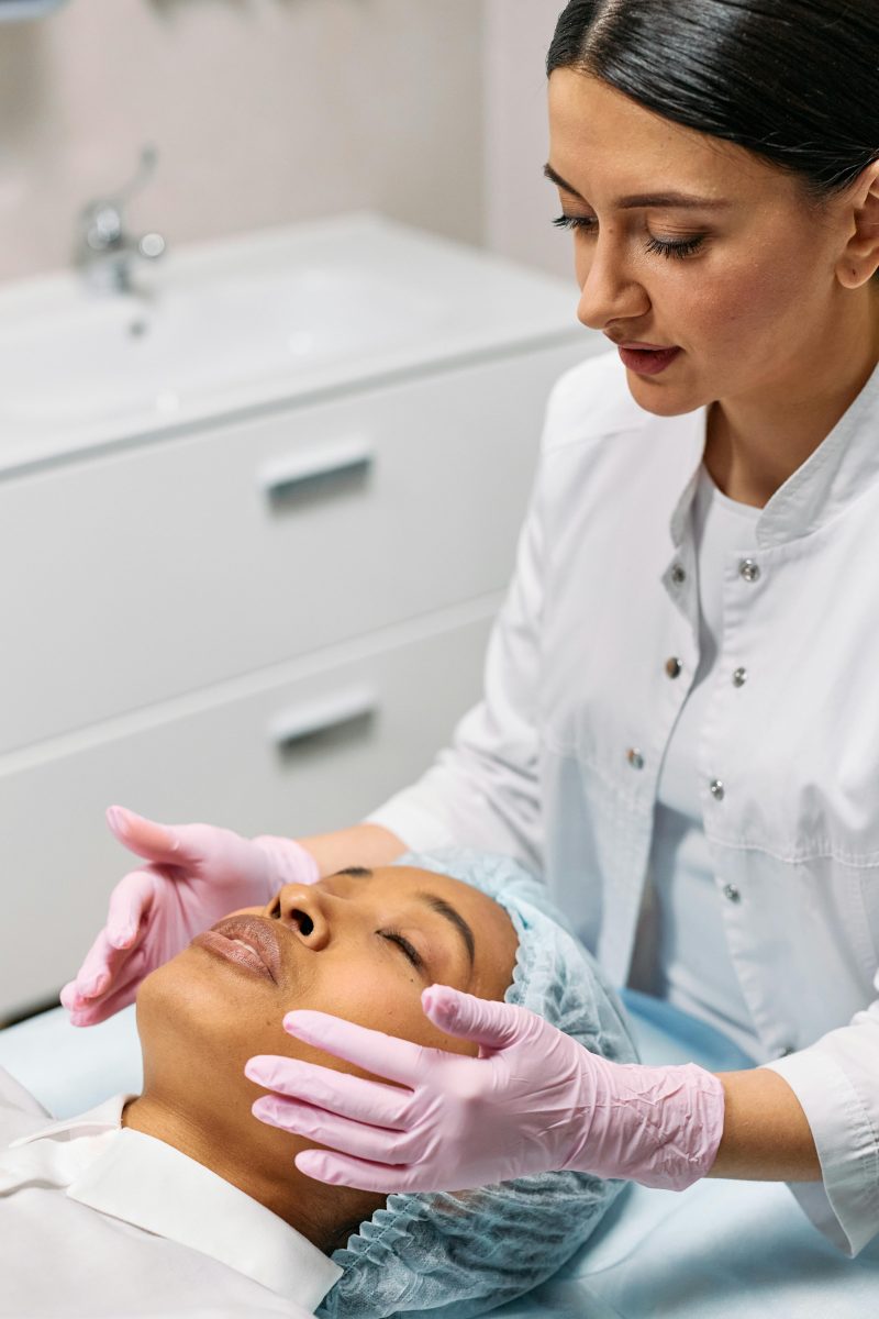 A skincare professional wearing pink gloves applying a wax cleaner to a client’s face after a waxing session.