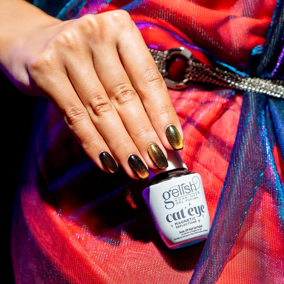 Hand with shimmering, multicolored cat eye nails holding a Gelish cat eye gel polish bottle. The bottle reads “Gelish CAT EYE MAGNETIC GEL POLISH.” The nails show a color shift from gold to green to brown. The vibrant background enhances the stylish look.