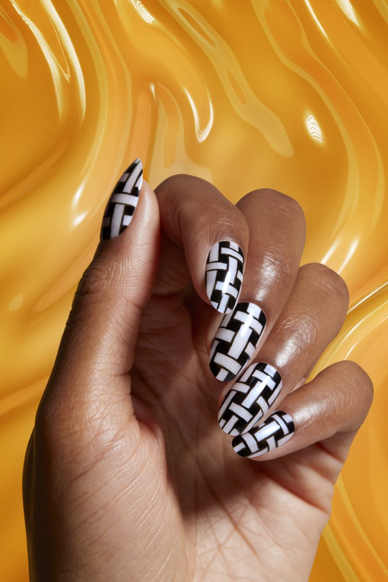 A close-up of a hand with nails painted in a black and white geometric pattern, demonstrating the long-lasting and glossy finish of OPI Infinite Shine New Formula. The background is a smooth, glossy yellow surface, highlighting the intricate nail art and vibrant polish quality.