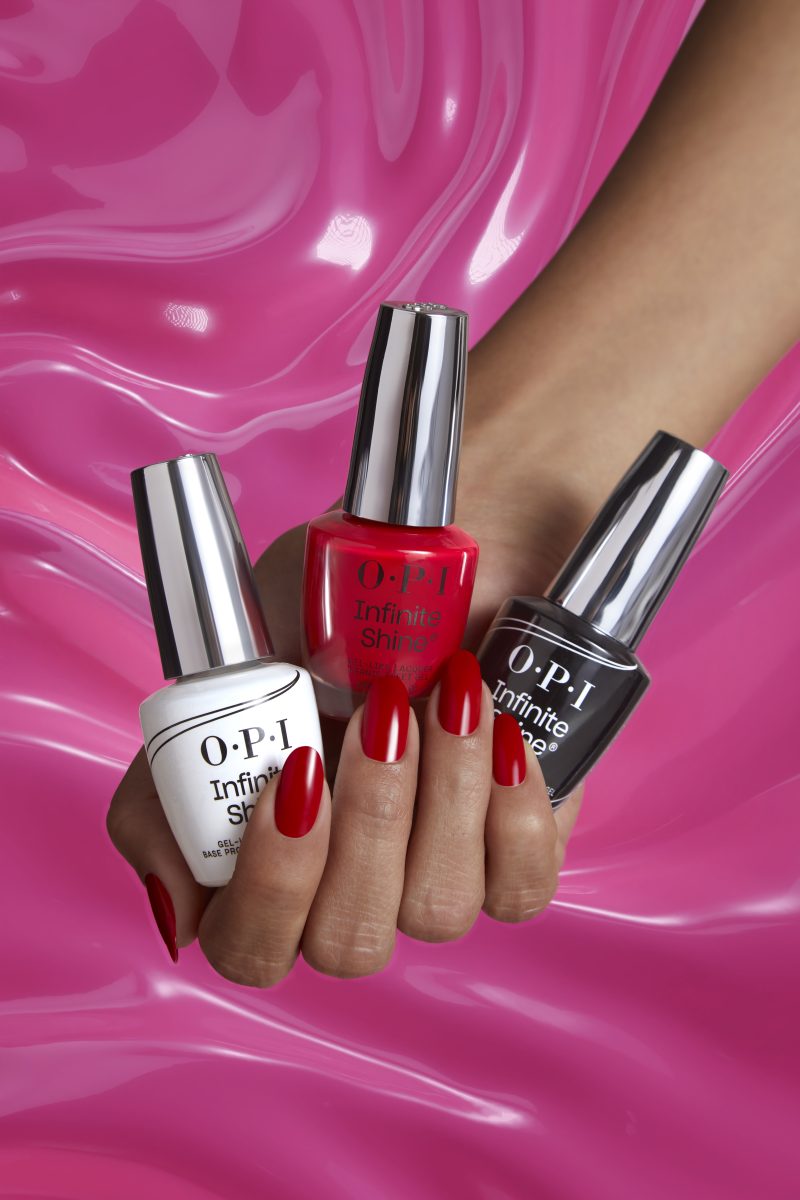 A hand with glossy red-painted nails holds three bottles of OPI Infinite Shine New Formula nail polish in white, red, and black colors against a vibrant pink wavy background. The image highlights the vibrant shades and high-gloss finish of the nail polish.